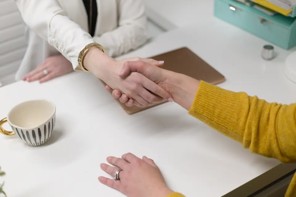 Dos personas se dan la mano por encima de una mesa de despacho, con una taza de café al lado.